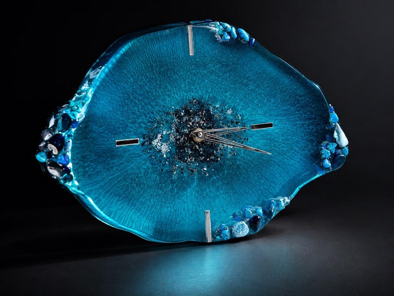 May include: An irregular, geode-inspired wall clock featuring a translucent, vibrant blue resin body. The center is filled with dark, shimmering crushed glass and metallic flakes. It includes silver-toned clock hands and three silver hour markers.