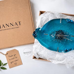 May include: A blue resin wall clock with a geode-inspired center, featuring silver hands and metallic accents. It is packaged in a cardboard box labeled MANNAT ARTISAN RESIN CREATIONS, accompanied by a thank you tag and a small circular brand sticker.