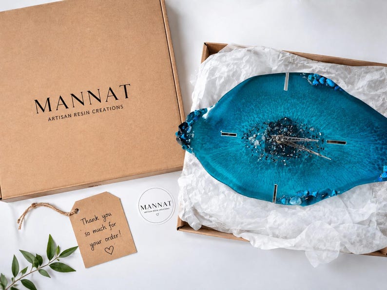 May include: A blue resin wall clock with a geode-inspired center, featuring silver hands and metallic accents. It is packaged in a cardboard box labeled MANNAT ARTISAN RESIN CREATIONS, accompanied by a thank you tag and a small circular brand sticker.