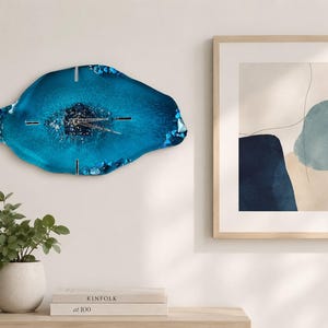 May include: An irregular, geode-inspired wall clock featuring a deep blue resin surface with a textured, crystalline center. The clock has silver-toned hands and thin, metallic hour markers positioned at the twelve, three, six, and nine o'clock positions.