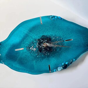 May include: A wall clock with an irregular, geode-inspired shape made of translucent teal resin. The center features dark, textured mineral accents and silver clock hands. The edges are decorated with small, polished blue and teal stones.