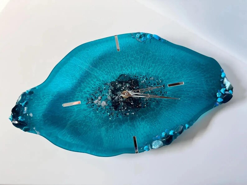 May include: A wall clock with an irregular, geode-inspired shape made of translucent teal resin. The center features dark, textured mineral accents and silver clock hands. The edges are decorated with small, polished blue and teal stones.