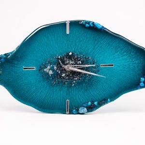 May include: An irregular, oval-shaped wall clock made of teal resin with a glossy finish. The center features a cluster of dark gray and silver metallic flakes. The edges are decorated with small blue and teal stones. It has silver clock hands.