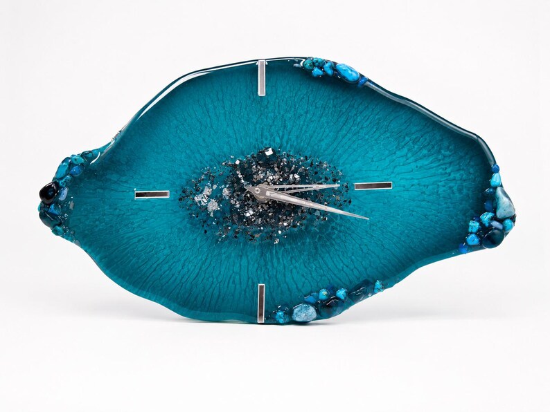 May include: An irregular, oval-shaped wall clock made of teal resin with a glossy finish. The center features a cluster of dark gray and silver metallic flakes. The edges are decorated with small blue and teal stones. It has silver clock hands.
