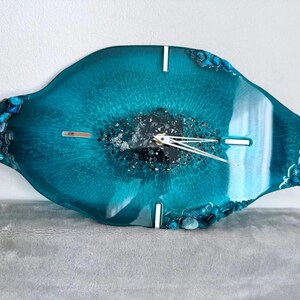 May include: An oval-shaped wall clock made of teal resin with a glossy finish. The center features dark, textured material with silver accents. It has silver clock hands and four silver rectangular markers at the twelve, three, six, and nine o'clock positions. The edges are decorated with small, dark blue and teal stones.