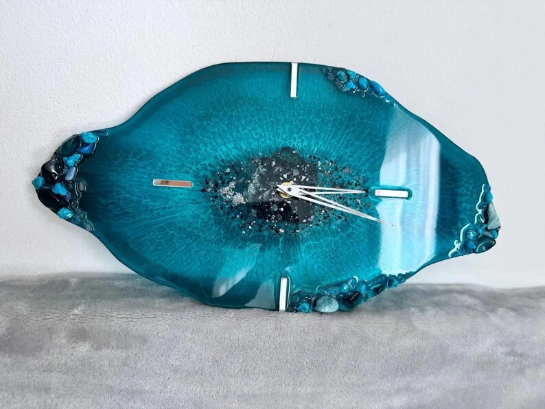 May include: An oval-shaped wall clock made of teal resin with a glossy finish. The center features dark, textured material with silver accents. It has silver clock hands and four silver rectangular markers at the twelve, three, six, and nine o'clock positions. The edges are decorated with small, dark blue and teal stones.