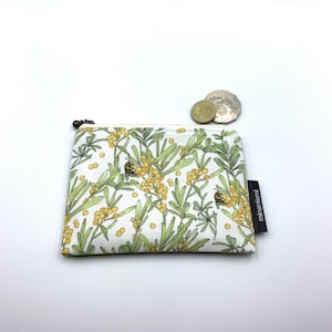 May include: A small, rectangular zipped pouch with a white background and a botanical pattern of green leaves and yellow berries. The pouch has a black zip pull and a small black tag that reads "minaniemi". Two coins are next to the pouch.