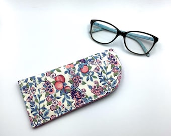 May include: A white and pink floral glasses case with a pink lining. The case has a curved top and a floral pattern with pink and blue flowers and green leaves. The case is lying on a white surface.