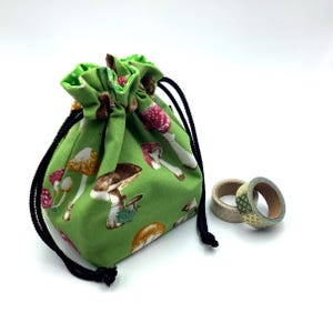 May include: Green drawstring bag with a mushroom print. The bag is made of fabric and has a black drawstring. Two rolls of washi tape are in the background.
