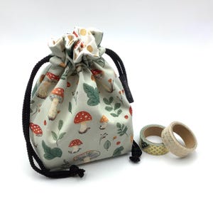 May include: A drawstring bag featuring a mushroom design in red, orange, and green on a pale green fabric. The bag has a black drawstring, and is next to three rolls of decorative tape.