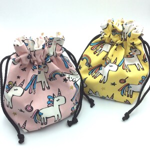 Könnte beinhalten: Zwei Kordelbeutel mit Einhorn-Designs. Ein Beutel ist rosa mit weißen Einhörnern und der andere ist gelb mit weißen Einhörnern.