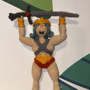 Puede incluir: Una figura de ganchillo hecha a mano de un guerrero, sosteniendo una espada gris por encima de la cabeza. La figura tiene un cuerpo color canela, cabello azul y lleva una corona dorada, pulseras y un atuendo rojo y dorado. Las botas son azules con ribete blanco.
