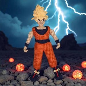 Puede incluir: Figura de ganchillo hecha a mano de un personaje con un atuendo naranja y negro con pelo amarillo puntiagudo. La figura está de pie sobre una superficie rocosa con orbes rojos brillantes. Un cielo azul con relámpagos en el fondo.