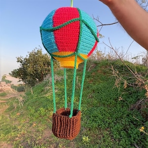 Könnte beinhalten: Ein handgehäkelter Heißluftballon in leuchtenden Farben. Der Ballon hat rote, blaue und gelbe Abschnitte, grüne Akzente und einen braunen Korb. Er hängt vor einem Hintergrund aus grünem Laub und blauem Himmel.