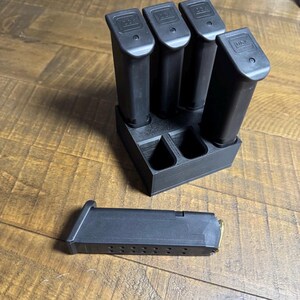 May include: A black plastic magazine storage rack holding three black Glock magazines in an upright position, with two empty slots visible. A single black Glock magazine with a visible brass cartridge is lying horizontally in front of the rack.
