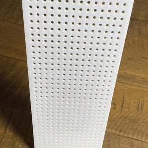 May include: A white rectangular display board featuring a grid of evenly spaced circular holes. The flat panel stands upright, providing a structured surface with numerous perforations designed for organizing or hanging various small items.