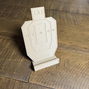 May include: A beige, textured, upright shooting target model featuring a central rectangular zone marked with the letter A, flanked by letters D, C, and C, D on the sides, with an additional A inside a small box at the top, all mounted on a base.