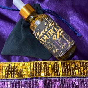 May include: A glass bottle with a dropper, filled with a golden liquid and herbs, labeled "Chewing Court Oil." The bottle rests on a purple cloth with a black pouch and gold and purple sequin trim. The label features scales of justice.