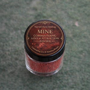 May include: A clear glass jar filled with red powder, topped with a black lid featuring a brown label. The label reads "MINE Commanding Love & Attraction Powder" with the text "Sacred Aura Healing" at the top.