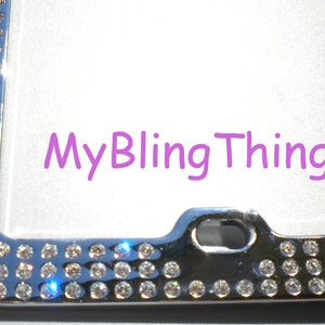 May include: A silver license plate frame with a row of clear rhinestones around the edge. The frame has a small hole in the top center for a license plate screw. The text "MyBlingThingz" is printed in purple on a white background.