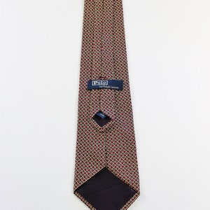 May include: A burgundy silk tie with a geometric pattern of small circles in gold and black. The tie has a navy blue label that reads "Polo by Ralph Lauren".