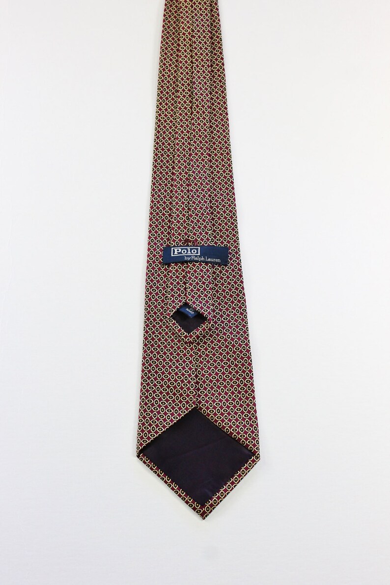 May include: A burgundy silk tie with a geometric pattern of small circles in gold and black. The tie has a navy blue label that reads "Polo by Ralph Lauren".