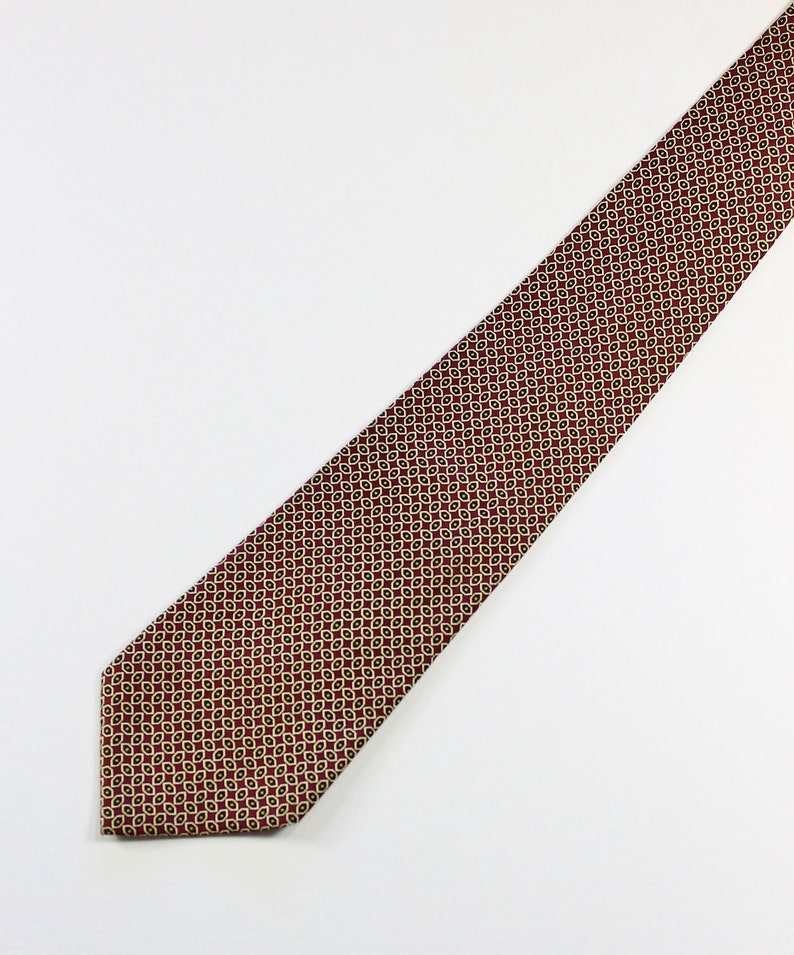 May include: A red patterned tie with a geometric design in gold. The tie is made of a smooth fabric.