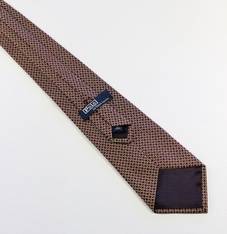 May include: A burgundy silk tie with a gold geometric pattern. The tie has a navy blue label that reads "Polo by Ralph Lauren".
