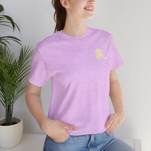 May include: A light purple t-shirt featuring a gold-colored outline of the state of Alaska on the left chest. The shirt is made of a soft material and has short sleeves. The person is wearing blue jeans.