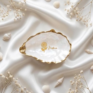 Peut inclure: Un vide-poche en forme de coquille d'huître blanche avec un bord peint en doré. L'intérieur présente un motif floral doré et le prénom Victoria écrit en lettres cursives. La coquille possède une finition nacrée et irisée sur sa surface.