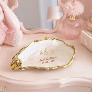 Könnte beinhalten: Eine weiße Austernschalen-Schale mit goldfarbenem Rand. In der Mitte befindet sich eine goldene Grafik mit zwei ineinander verschlungenen Ringen über dem Text "Jack & Annie" und dem Datum "08.08.2026" in goldener Schrift.