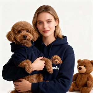 以下が含まれることがあります： 女性と小型犬が、お揃いのネイビーブルーのパーカーを着ています。犬は女性の腕の中にいます。女性のパーカーには犬の顔のグラフィックと、下に「arlie」という名前が刺繍されています。テディベアが背景にあります。