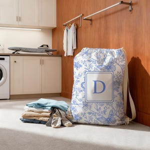 Op de afbeelding: Een waszak met een blauw en wit vlinder- en bloemenpatroon. De tas heeft een trekkoordsluiting en een grote letter "D" in een vierkant kader. Een stapel opgevouwen kleding en sokken staat voor de tas.