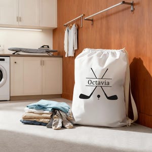 Puede incluir: Una bolsa de lavandería blanca con cordón, con un diseño de palos de golf negros y el nombre "Octavia". La bolsa está en un lavadero con ropa doblada y una lavadora.