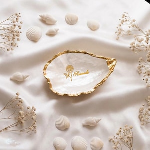 Può includere: Un piattino a forma di conchiglia di ostrica bianca con bordo smerlato dipinto in oro. L'interno presenta una grafica dorata di una rosa e il nome Amanda scritto in corsivo. La conchiglia è circondata da piccole conchiglie bianche e fiori.