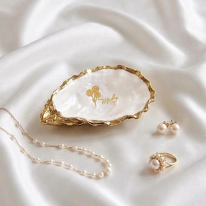 May include: A white oyster shell trinket dish with a gold-painted floral design and the script text "Marley" in the centre. The dish is accompanied by a delicate pearl necklace, a pair of pearl stud earrings, and a gold pearl-encrusted ring.
