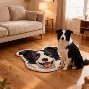 May include: A black and white Border Collie dog sitting next to a matching dog-face rug. The rug is shaped like a smiling dog's face with brown eyes and a pink tongue. The dog is on a hardwood floor in a living room setting.