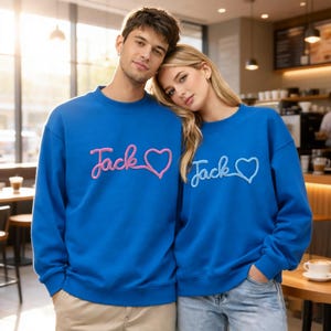 Könnte beinhalten: Zwei Personen tragen passende blaue Sweatshirts. Jedes Sweatshirt hat den Namen "Jack" in Schreibschrift gestickt, verbunden mit einem Herzsymbol. Eines hat rosa Stickerei, das andere hellblau.