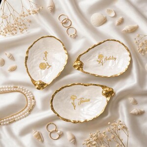 Könnte beinhalten: Drei weiße Austernschalen-Schmuckschalen mit goldfarbenen Rändern. Jede Schale zeigt eine goldene Blumenillustration und einen Namen in Schreibschrift: Emily mit einer Gänseblümchen, Ashley mit einer Rose und Jessica mit einer Nelke. Die Schalen liegen auf Stoff.