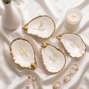 May include: Four white oyster shell trinket dishes with gold-painted edges. Each dish features custom gold lettering and graphics, including names like Stella & Jackson or Jack & Annie, dates, and wedding ring icons. The shells have a glossy, textured finish.