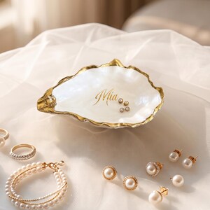 Puede incluir: Un plato decorativo en forma de concha de ostra blanca con borde dorado y el nombre Mia impreso en letras doradas en el centro. El plato contiene pequeños pendientes de perlas, rodeado de otras joyas de perlas y anillos sobre tela blanca.