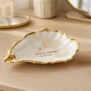 May include: A decorative oyster shell trinket dish with a pearlescent white interior and a metallic gold-painted rim. The centre features gold lettering that reads S J, Stella & Jackson, and the date 06.06.2026, accompanied by a small floral graphic.