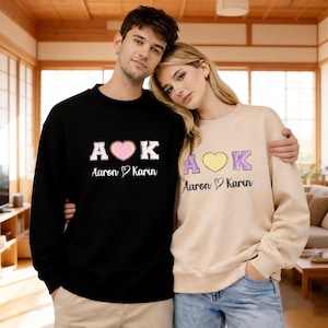 May include: Two people wearing matching sweatshirts. One is black with "A♥K" in white, a pink heart, and the names "Aaron & Karin". The other is beige with the same design, but with a yellow heart. The text is embroidered.