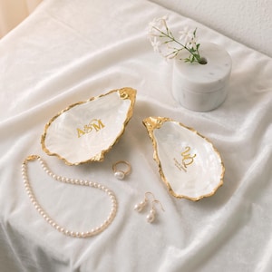 May include: Two white oyster shell dishes with gold-painted edges and gold lettering. One dish features the letters A&M, the other displays W&Q with text. A pearl necklace, a pearl ring, and pearl earrings are arranged alongside a white marble vase.