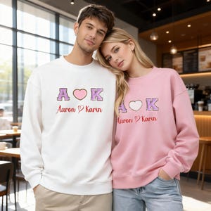 May include: Two people wearing matching sweatshirts. One is white, the other is pink. Both sweatshirts feature the letters "A", a heart, and "K" embroidered in purple and pink, with the names "Aaron" and "Karin" underneath.