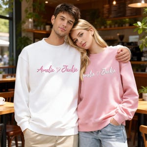 Peut inclure: Deux personnes portant des sweat-shirts assortis. L'un est blanc, l'autre rose. Les deux sweat-shirts portent les noms "Amelie" et "Jackie" brodés en cursive, avec un symbole de cœur entre les noms.