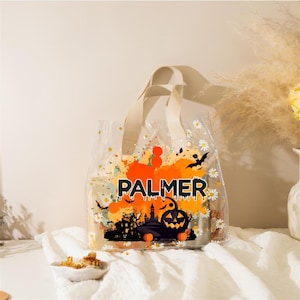 Puede incluir: Bolso tote transparente con asas color crema y un diseño de Halloween. El diseño incluye el nombre "PALMER" en naranja, una casa embrujada, murciélagos y calabazas. La bolsa está decorada con pequeñas flores blancas.