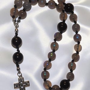 May include: A rosary featuring translucent grey beads and solid black beads strung on black cord. The necklace includes a silver-toned metal cross pendant with an intricate openwork filigree design, connected by a silver-toned metal link.