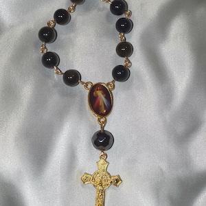 May include: A rosary featuring dark, round beads connected by gold-colored links. The rosary includes a gold-colored cross and a central medallion with a religious image. The beads appear to be made of a dark, possibly obsidian-like material.