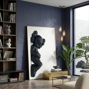 May include: A large framed artwork featuring a black sculpture of a figure with crossed-out eyes and a rounded head. The artwork is mounted on a white background with a gold frame. The room has a modern design with a dark blue wall and a wooden bookshelf.